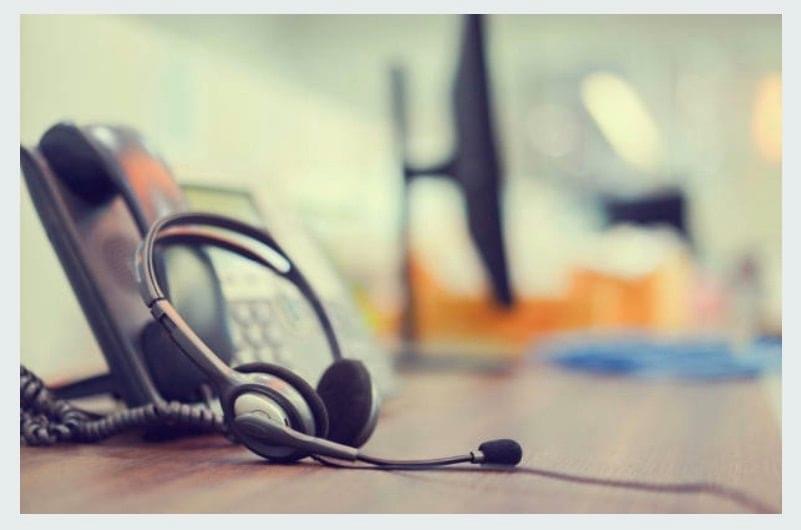 A close-up image of a headset with a microphone placed on a desk. In the background, there is a blurred office phone and a computer monitor. The image suggests a work environment, likely a call center or an office setting where communication tools are essential. The focus on the headset highlights the importance of verbal communication in this workspace.